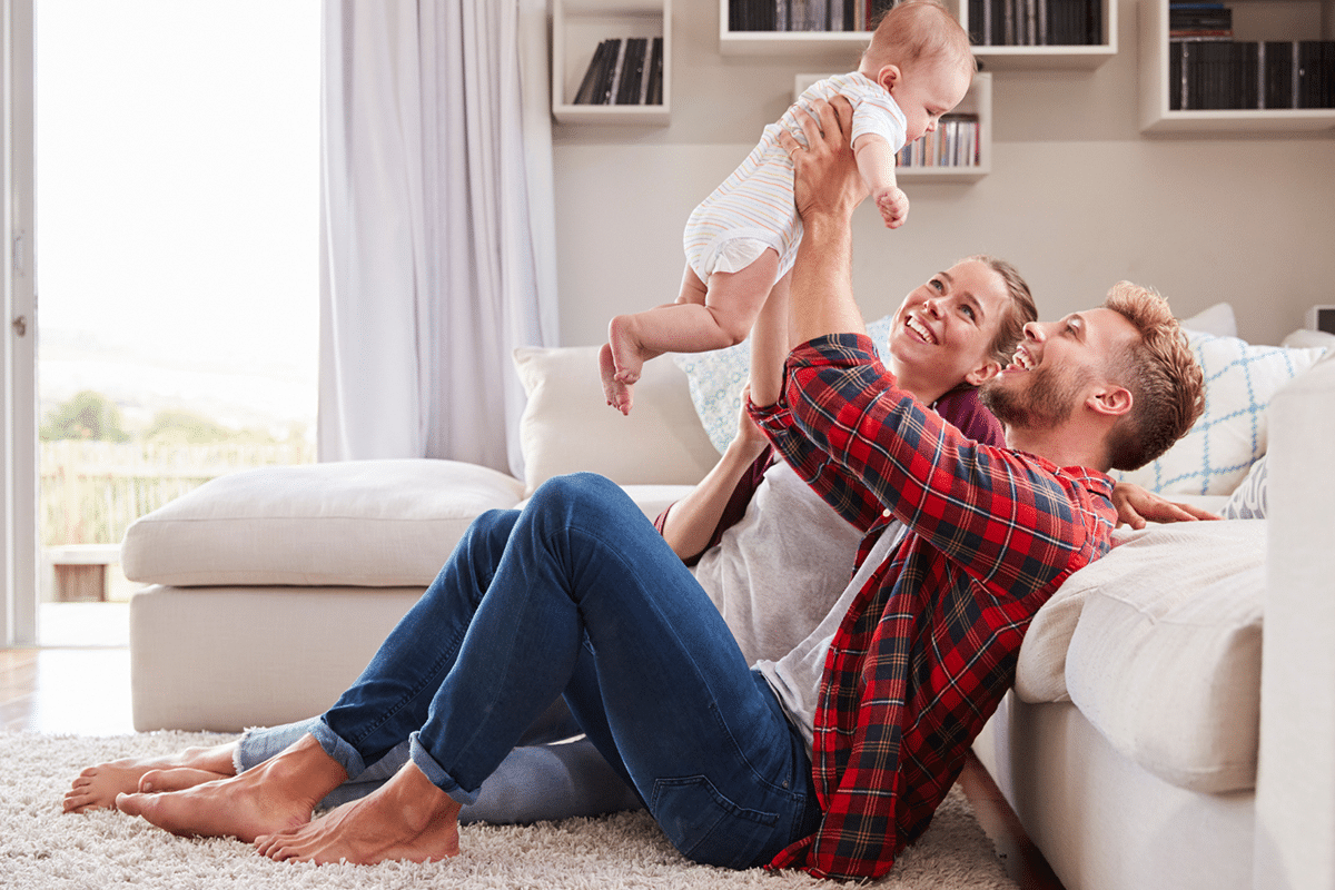 Two young smiling parents sitting on the floor in front of a couch looking up as they hold their baby in the air.